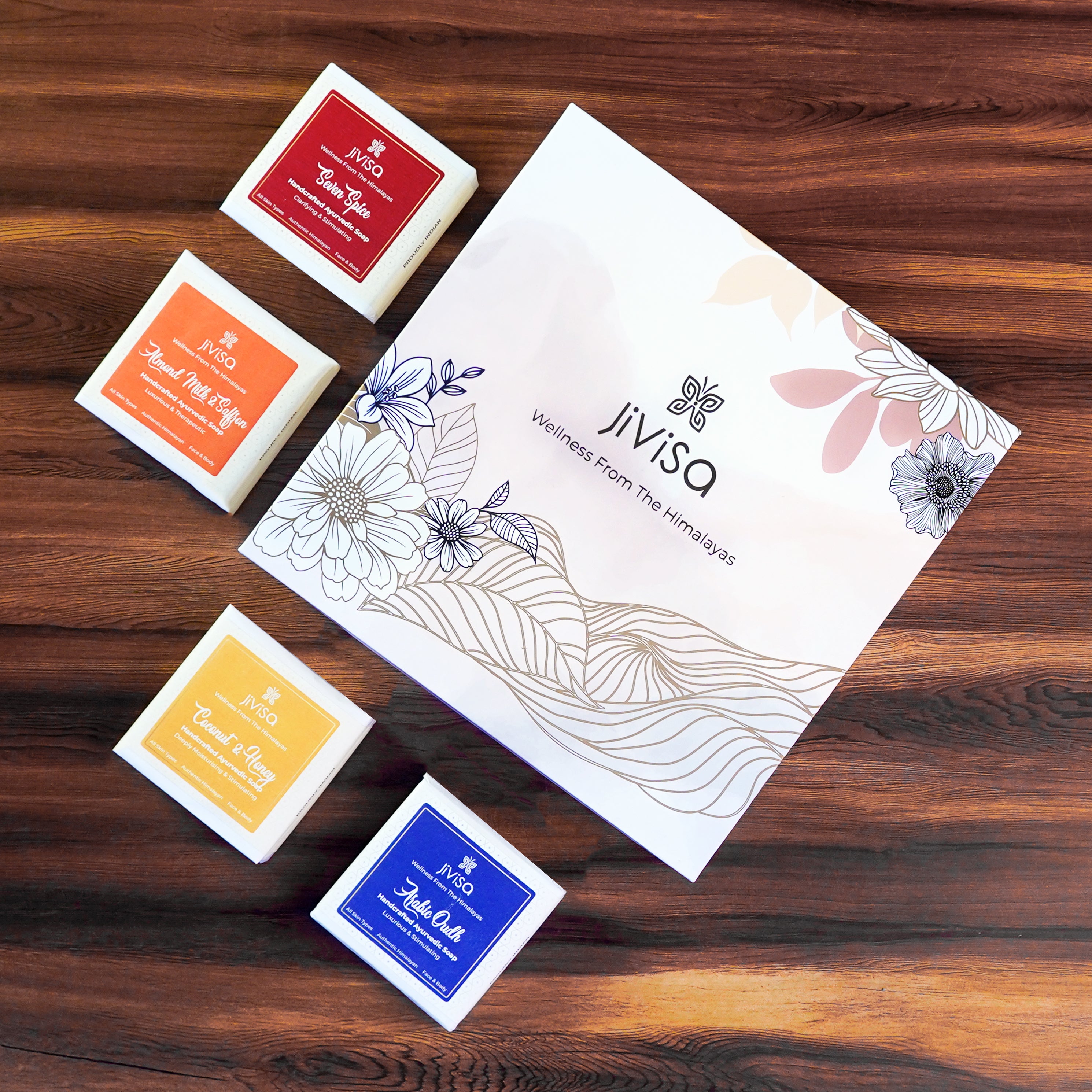 JiViSa Luxury Ayurvedic Handmade Soaps Gift Box. JiViSa four distinct Handmade soaps: Seven Spices, Arabic Oudh, Coconut &amp; Honey and Almond Milk &amp; Saffron are placed on the wooden surface around the box.