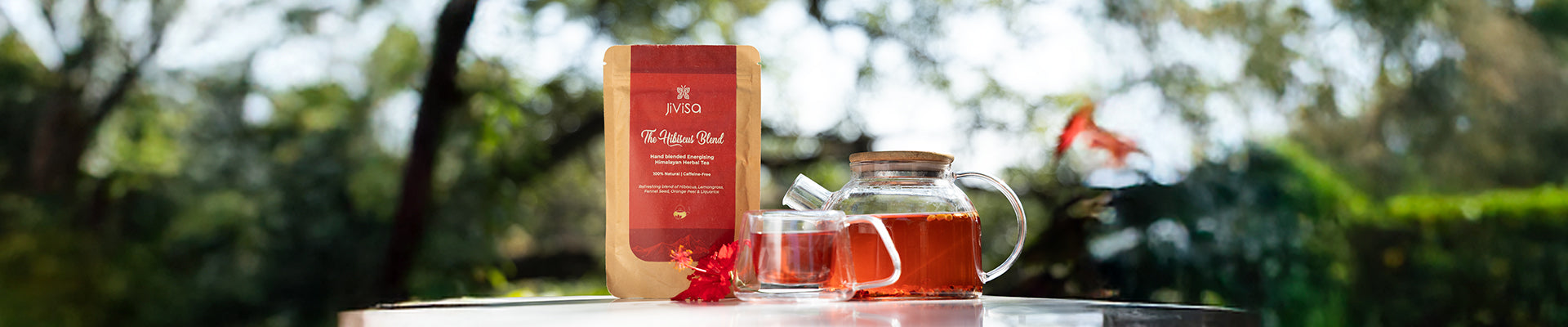 JiViSa Hibiscus Blend herbal tea pouch placed beside a glass teapot and cup filled with vibrant red hibiscus tea set in a calm outdoor setting with soft sunlight and greenery capturing the soothing antioxidant-refreshing caffeine-free essence of the blend that helps boost energy support digestion and promote glowing skin.