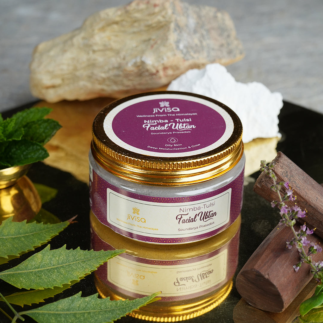 Image showcases a jar of JiViSa-Nimba Tulsi Facial Ubtan. The product is surrounded by its natural ingredients: fresh green neem leaves, sprigs of Tulsi with small purple flowers, sandalwood pieces, and what appears to be Fuller&
