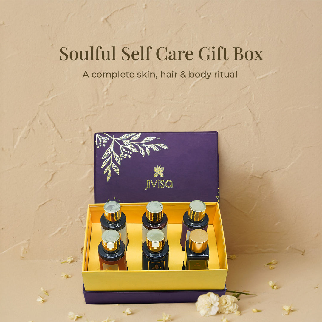 An elegant open JiViSa Soulful Self Care Gift Box on a textured beige background, with white petals scattered around. The deep purple lid, adorned with a gold floral design, is propped open, revealing the yellow tray holding six bestselling JiViSa&