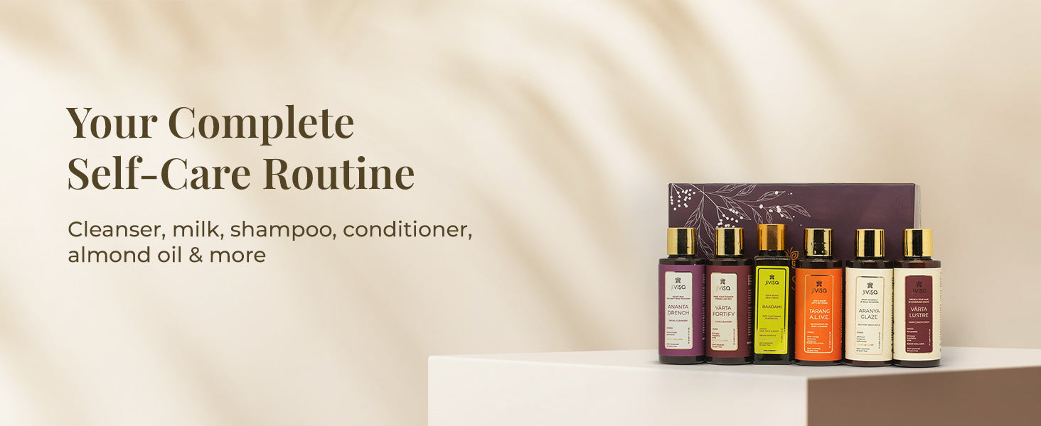 Six bestselling JiViSa's skin, hair and body essential bottles- Ananta drench face cleanser, Varta fortify hair cleanser, Badaami cold pressed oil, Tarang glaze body cleanser, Aranya glaze body milk and Varta lustre hair conditioner are lined up on a simple white pedestal against cream background with soft shadows. The tagline on the image says, "Your Complete Self-Care Routine," with a sub-headline listing some of the contents: "Cleanser, milk, shampoo, conditioner, almond oil & more.