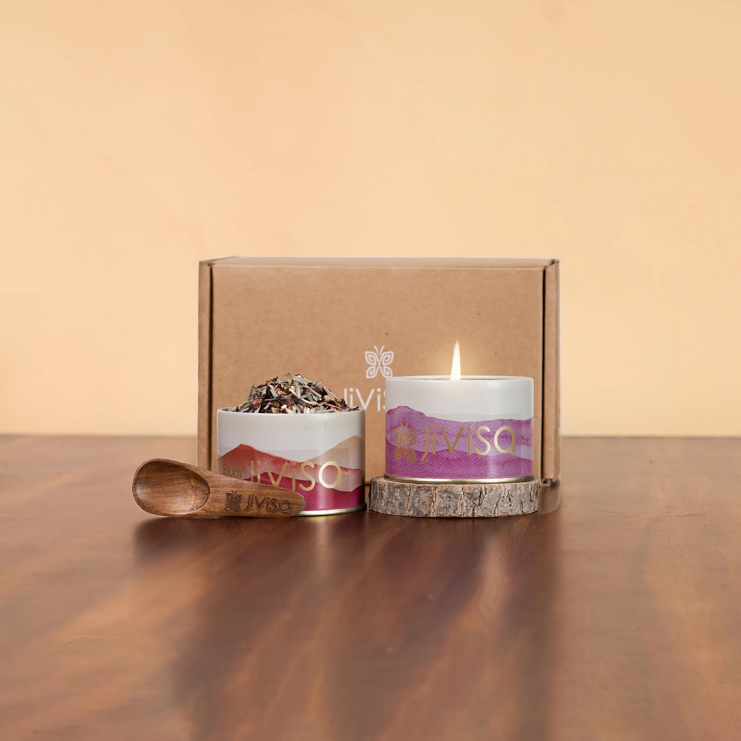 JiViSa herbal tea tin and lit soy candle with mountain-print labels displayed beside kraft gift box and wooden spoon on wood table