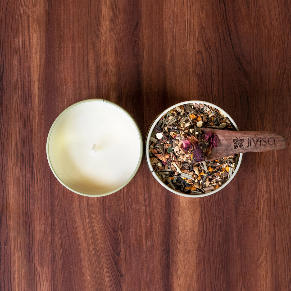 Overhead view of open JiViSa candle tin and loose-leaf herbal tea tin with rose petals, scooped by a wooden spoon on dark wood table
