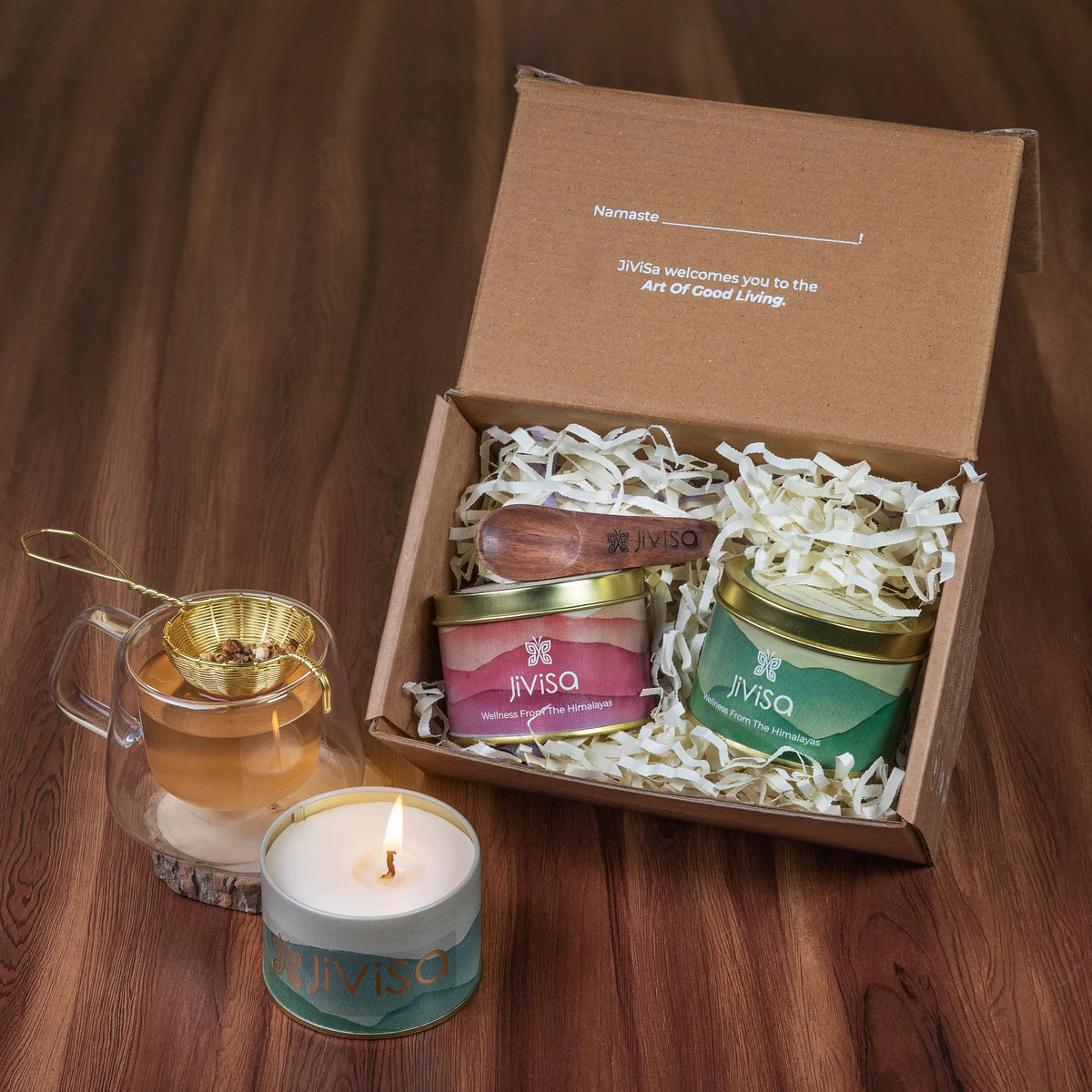 Open JiViSa gift box with Namaste welcome note, wooden spoon, mountain-print tea and candle tins, brewed herbal tea, and lit candle on wooden table