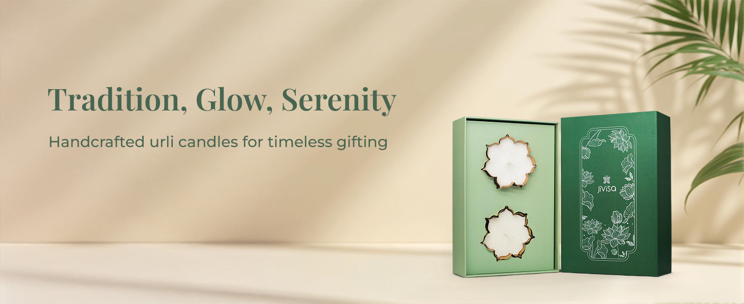An open JiViSa Urli Elegance Aroma gift box displayed on a light-colored surface with a plant in the background. The green box contains two handcrafted white JiViSa urli candles. On the left, the tagline reads "Tradition, Glow, Serenity", followed by text ‘Handcrafted urli candles for timeless gifting.’