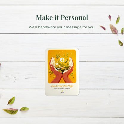 &quot;Make it personal – We’ll handwrite your message for you.&quot; A handwritten card with warm yellow and orange tones shows two hands holding blooming herbs with a glowing sun. Placed on a white surface, the image emphasizes the personal, heartfelt gifting experience JiViSa offers.