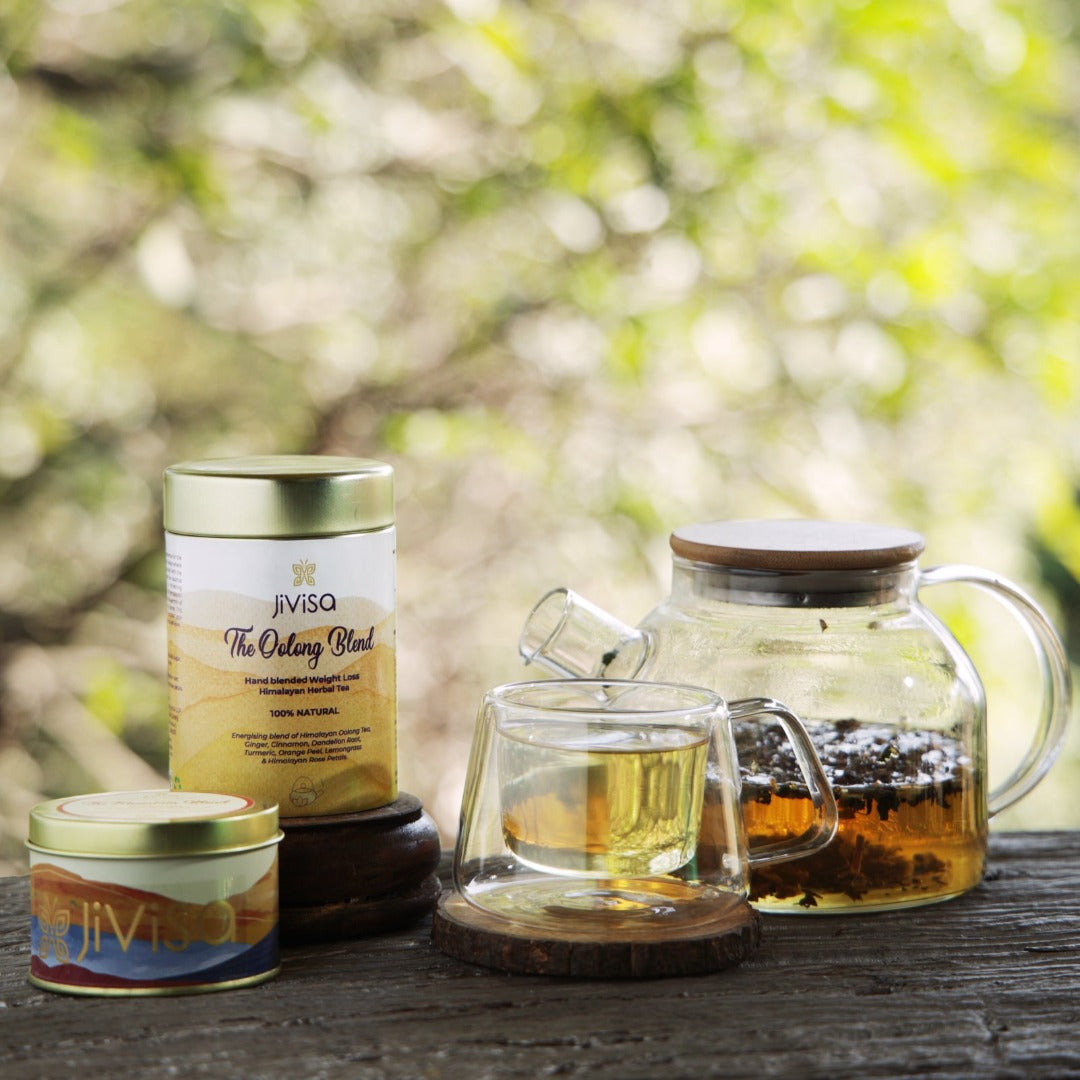 The image showcases 50gm of JiViSa-The Oolong Blend and 25gm of JiViSa-The Oolong Blend Weight loss Himalayan Herbal tea along with a clear glass teapot with a wooden lid contains steeped tea leaves, in front of the teapot, a clear double-walled glass mug rests on a small wooden coaster, filled with a light-colored Oolong Blend tea. The overall scene suggests a focus on natural, hand-blended teas and the ritual of tea preparation and enjoyment.