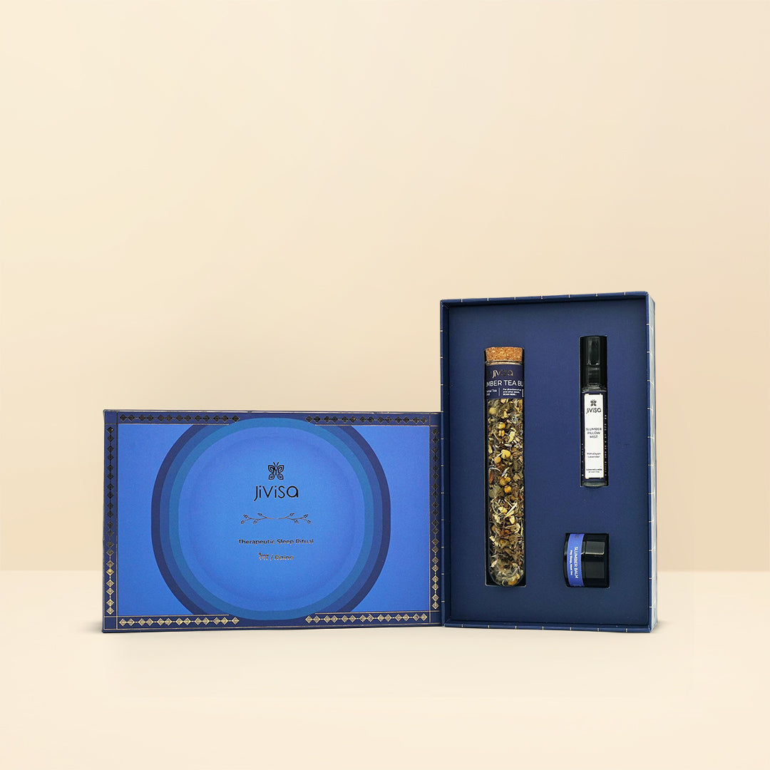 An elegant, open JiViSa Raina Sleep Ritual Kit gift box is presented on a simple, light beige background. The main box interior, which is standing upright, holds three items nestled in matching blue cutouts: a tall, clear glass tube of loose herbal tea (the Slumber Blend), a slender black roller bottle labeled "Raina Slumber" (the Pillow Mist), and a small, round blue jar (the Slumber Balm). The box features the JiViSa logo and circular design elements on the front flap. 