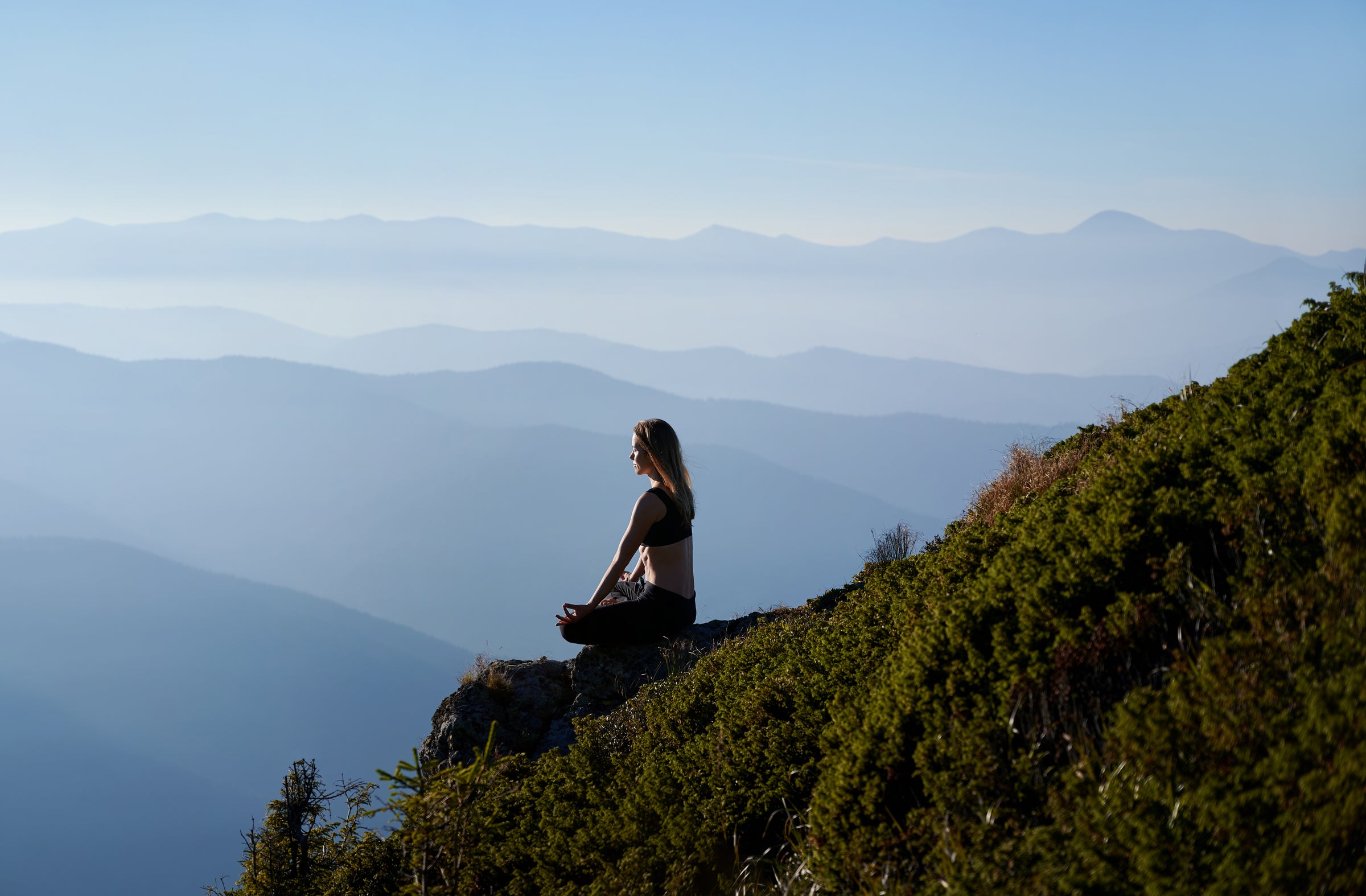 Woman meditating on a mountain peak surrounded by serene blue Himalayan ranges symbolizing JiViSa’s connection to mindfulness natural balance and the healing power of Ayurveda inspired living.