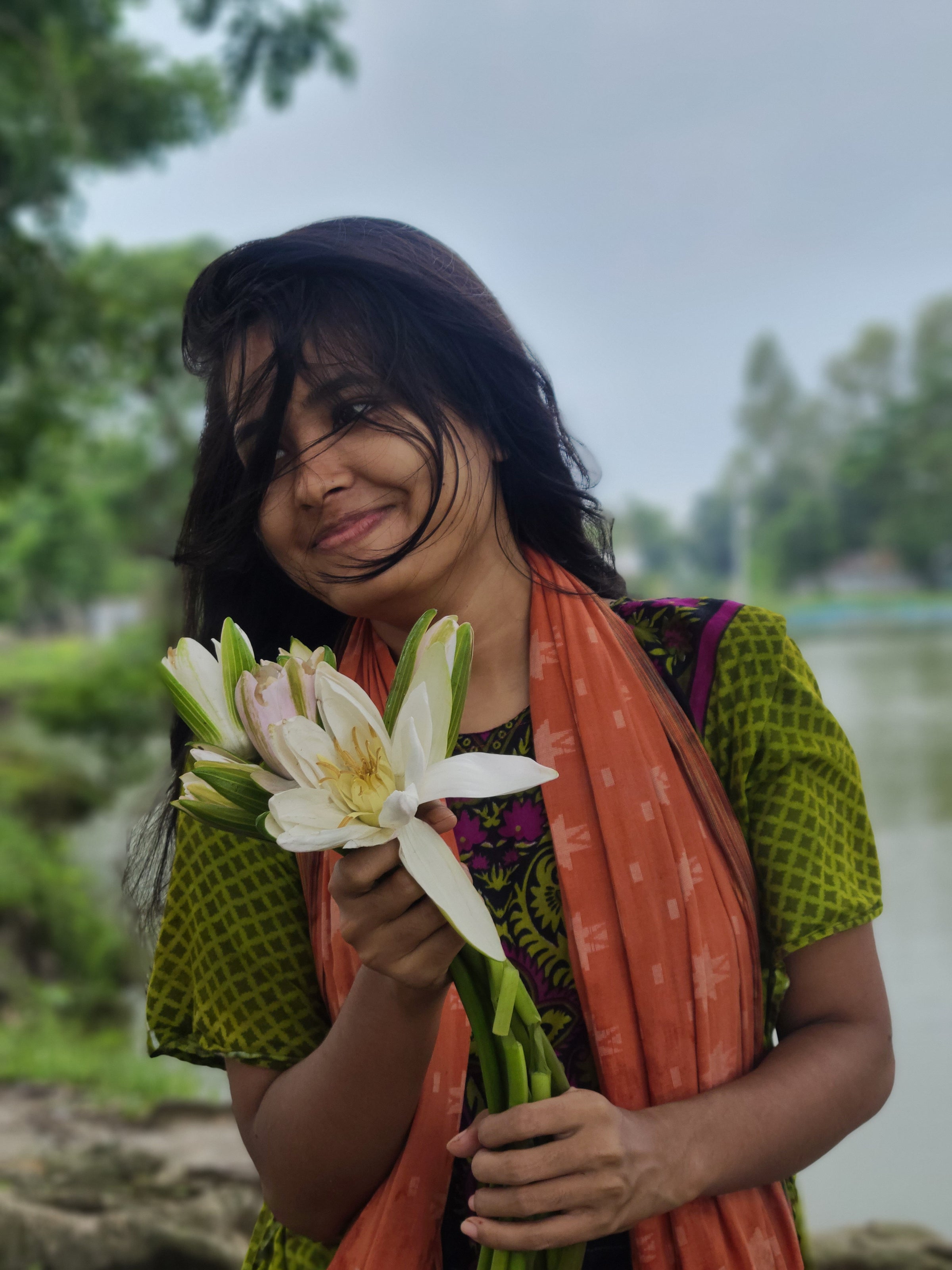 Smiling woman holding fresh white lotus flowers near a calm riverside reflecting JiViSa’s philosophy of natural beauty purity and joyful living inspired by Indian traditions and Ayurvedic wisdom.