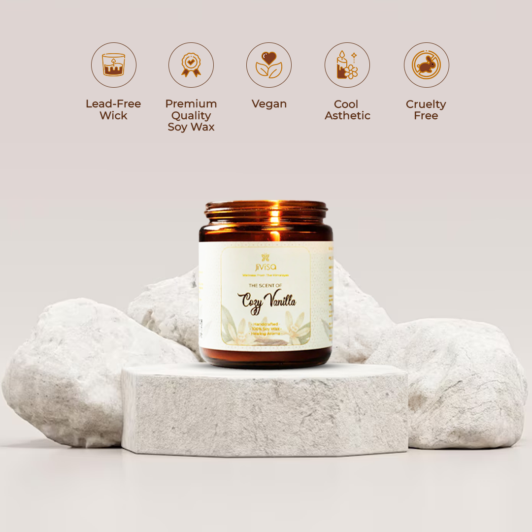 JiViSa Cozy Vanilla candle amber glass jar is displayed on a minimalist stone platform surrounded by soft white rocks, with five icons above it. The icons and accompanying text highlight key features: &quot;Lead-Free Wick,&quot; &quot;Premium Quality Soy Wax,&quot; &quot;Vegan,&quot; &quot;Cool Aesthetic,&quot; and &quot;Cruelty Free.&quot; The candle label reads: &quot;The Scent of Cozy Vanilla – Handcrafted • 100% Soy Wax • Healing Aroma.&quot;