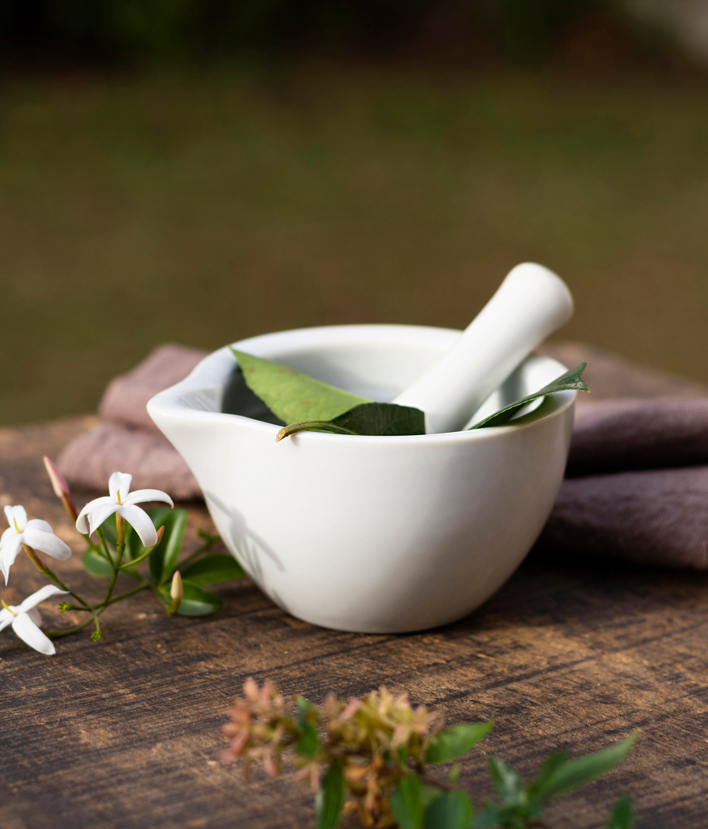 White ceramic mortar and pestle with green Ayurvedic leaves and jasmine flowers on a wooden surface symbolizing JiViSa’s blend of nature, purity, and science-backed skincare.